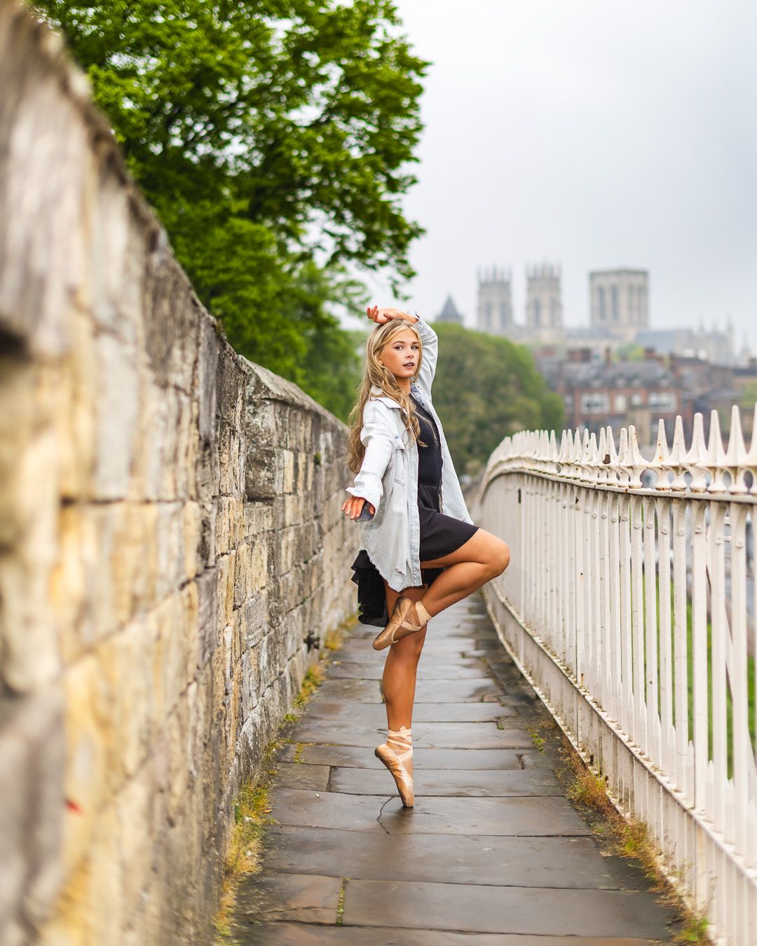 A ballet dancer poses en pointe on a stone walkway with York Minster visible in the background.