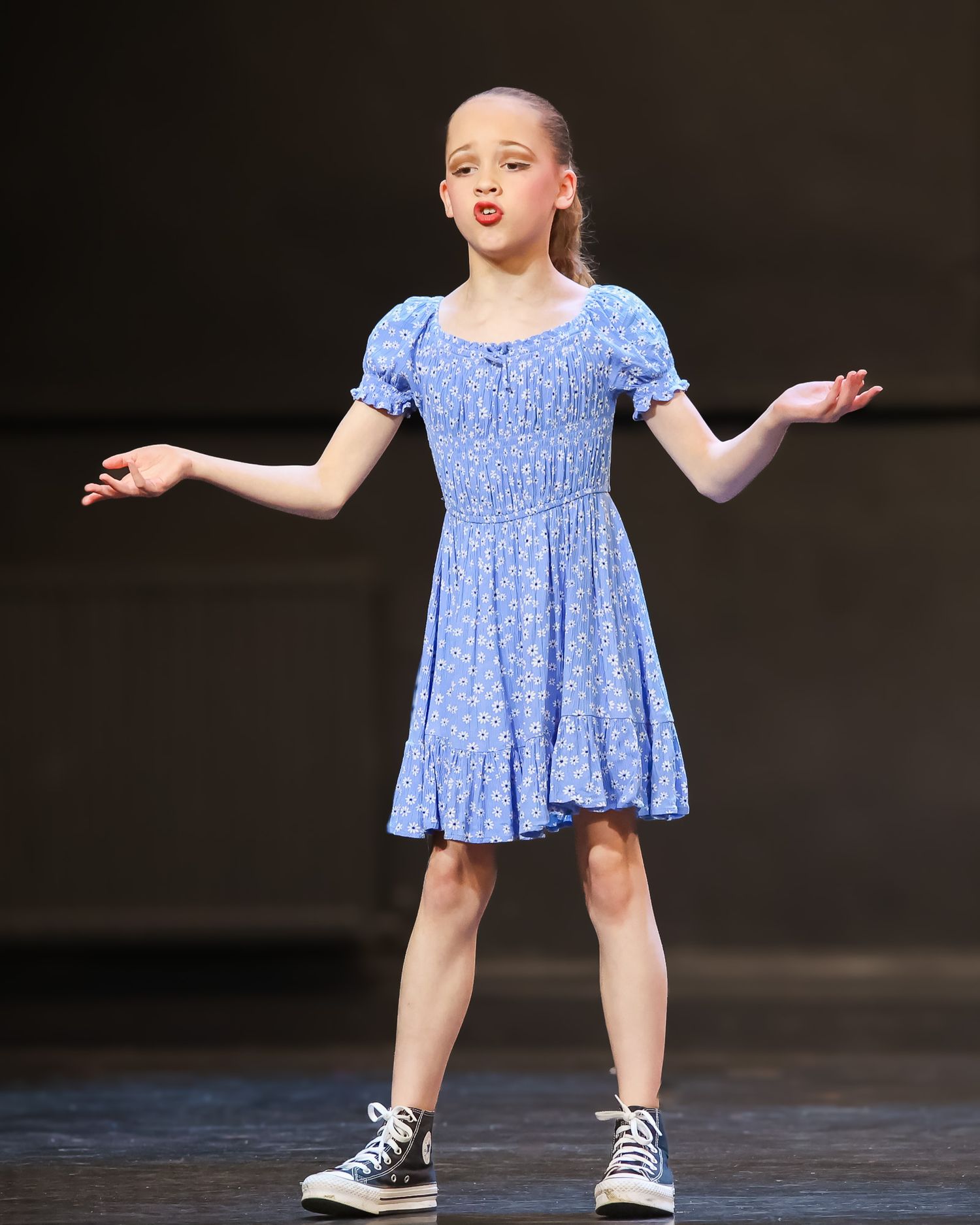 Young girl in blue floral dress and sneakers performing on stage with arms outstretched.