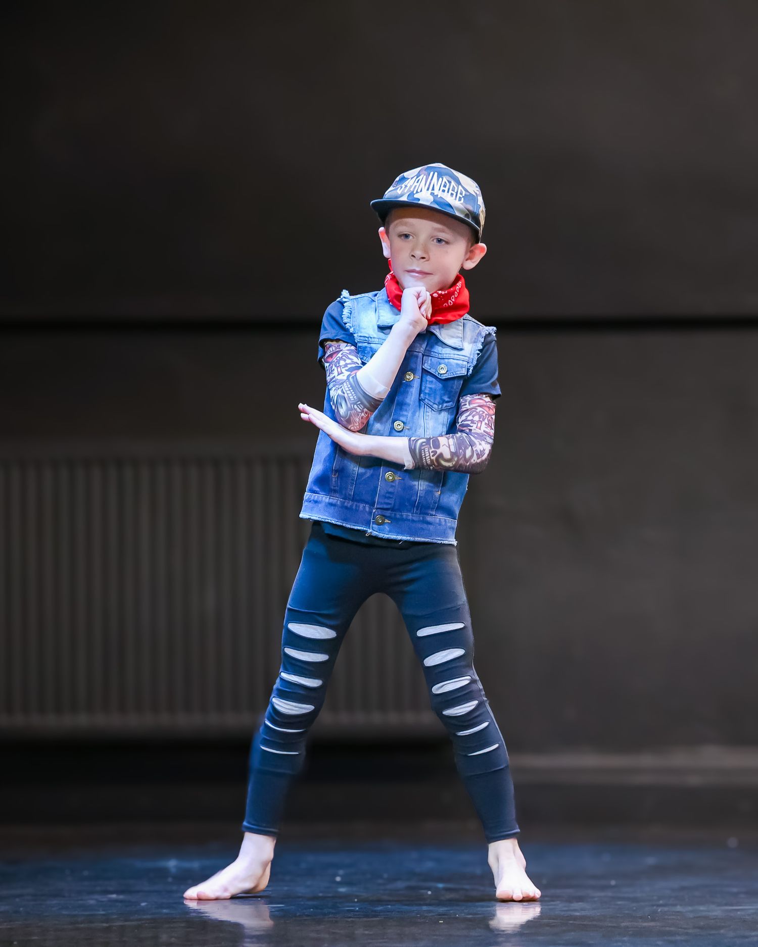 Young red-haired boy performing hip-hop dance on stage wearing denim vest, cap, and striped leggings.