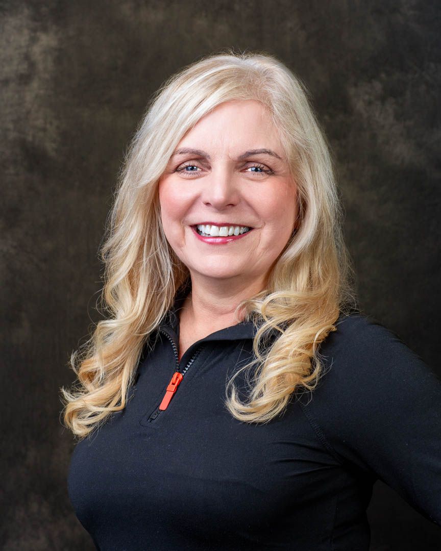Professional headshot of a smiling blonde woman with curly hair wearing a black zip-up jacket.