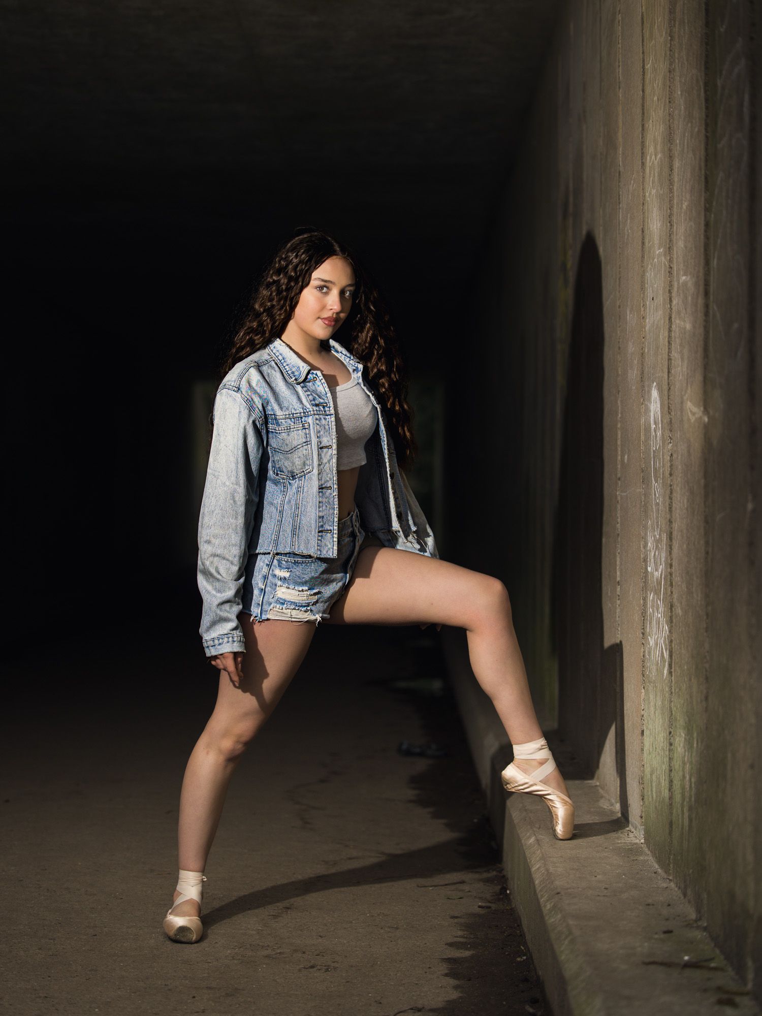 A dancer in a denim jacket and ballet shoes poses gracefully in a dark concrete hallway.