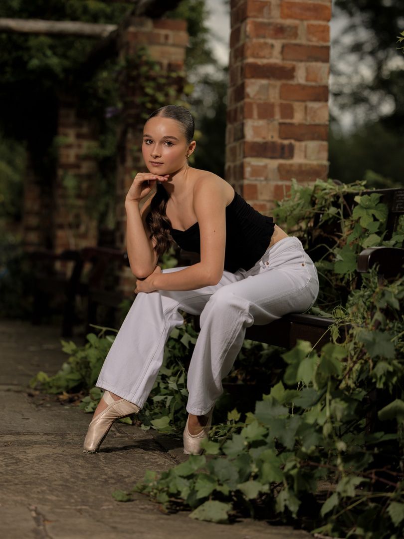 A dancer in white pants rests thoughtfully against a brick wall surrounded by green foliage.