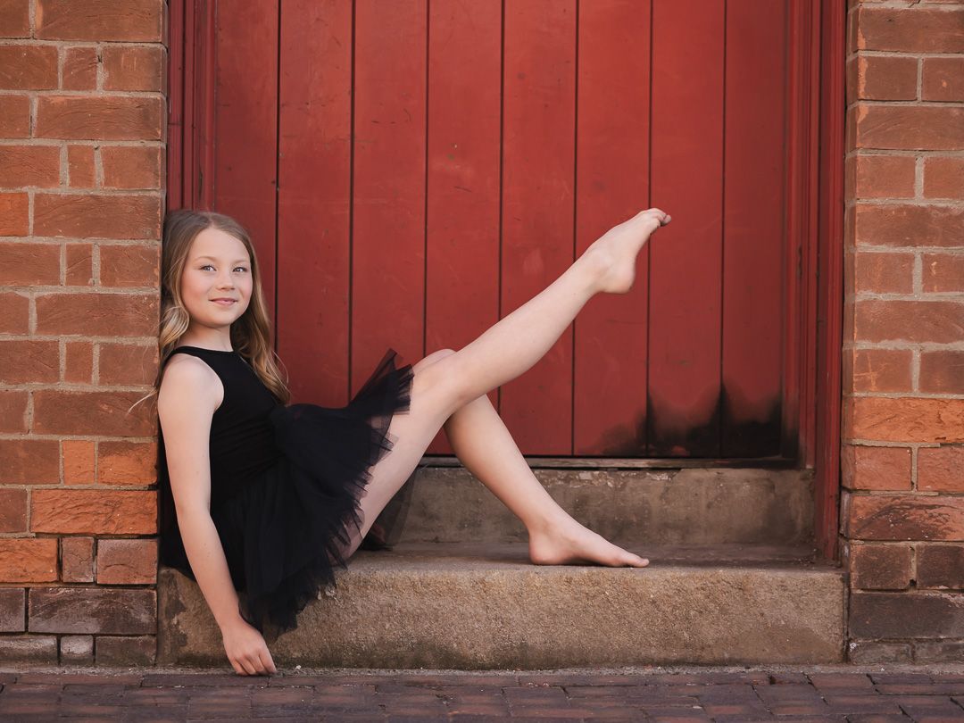 Dancer in black leotard and tutu poses gracefully against red wooden door and brick wall.