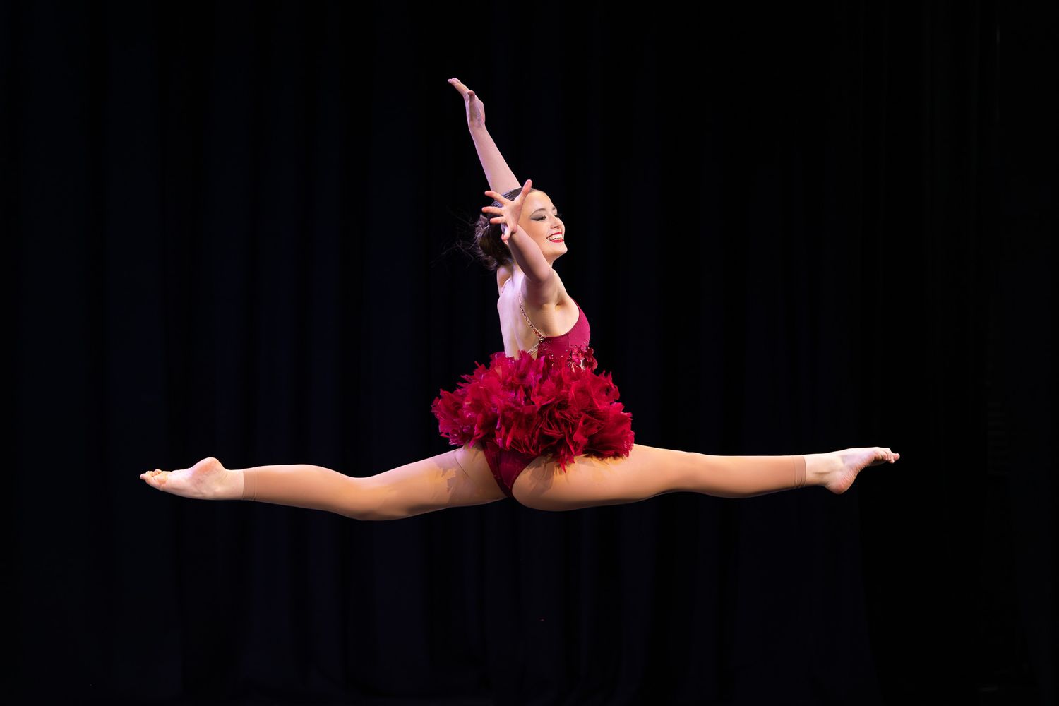 Dancer in red feathered costume performs a grand jeté split leap against a dark stage background.