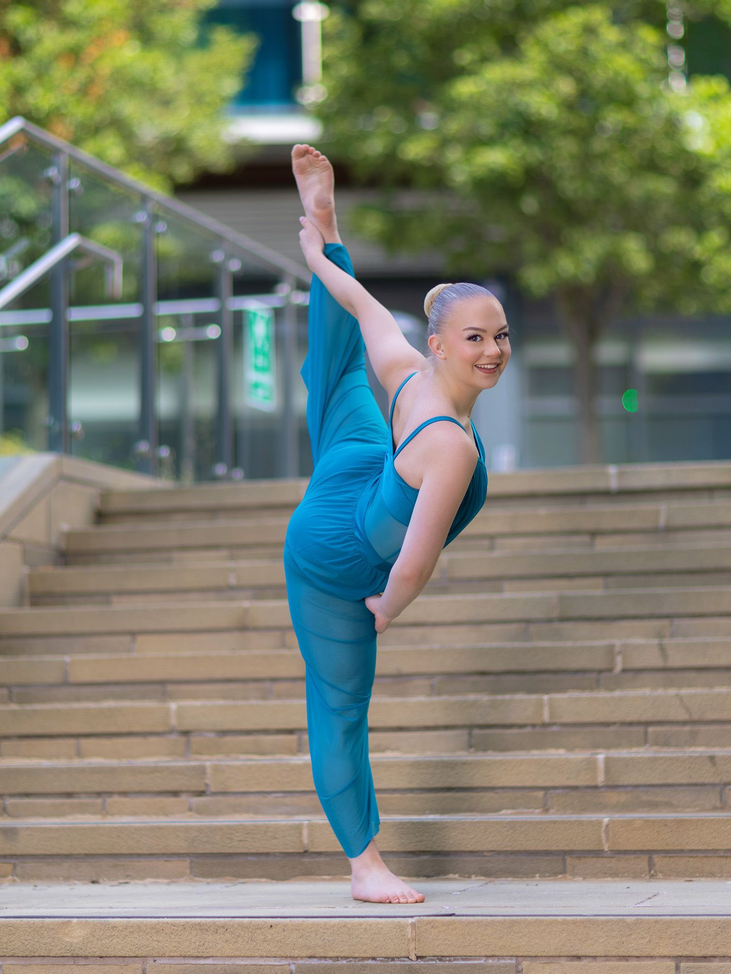 Flexible dancer in teal jumpsuit performs a standing split on outdoor steps surrounded by green trees.