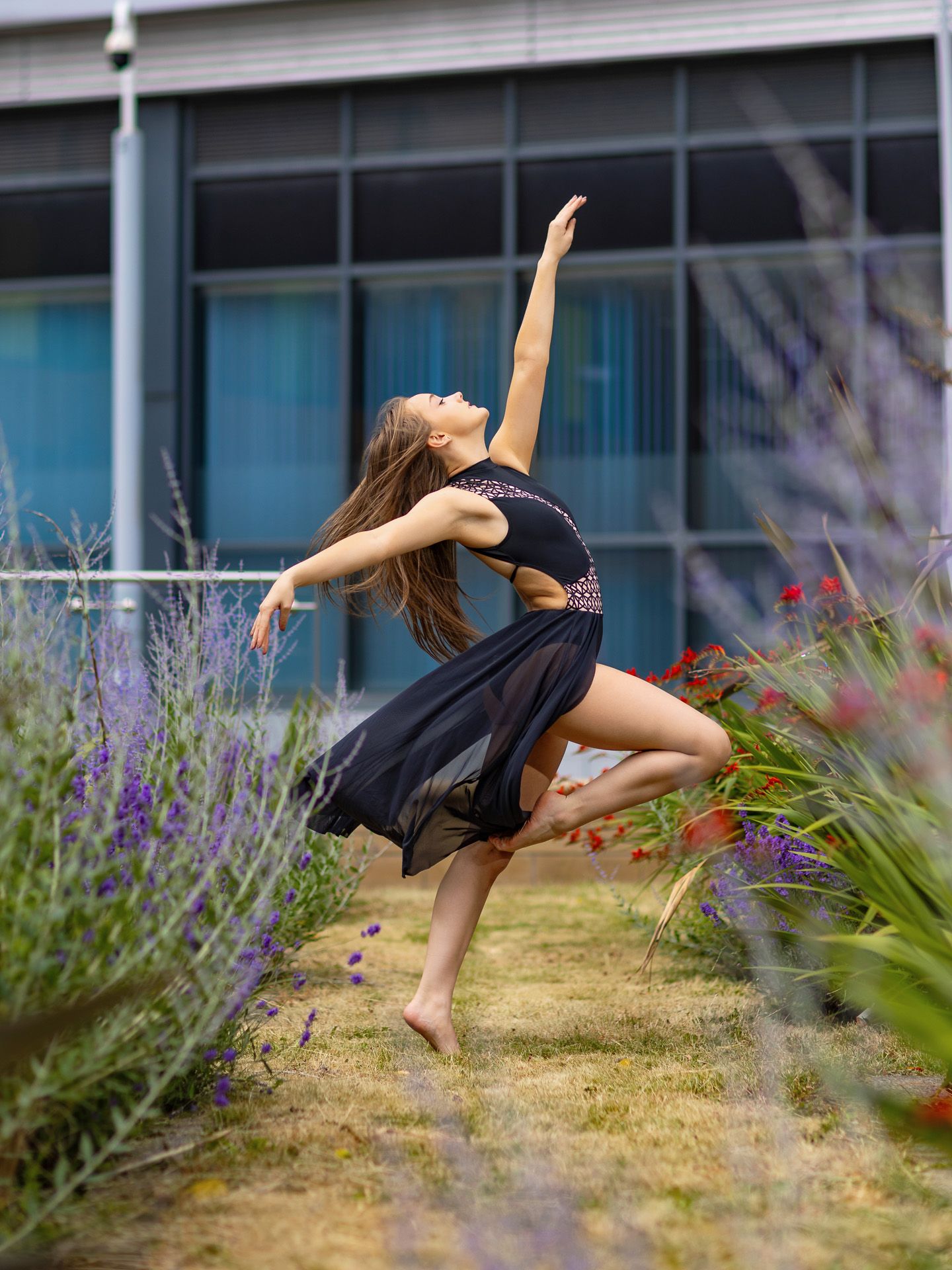A female dancer leaps gracefully in a black dress among colorful garden flowers outside a modern building.