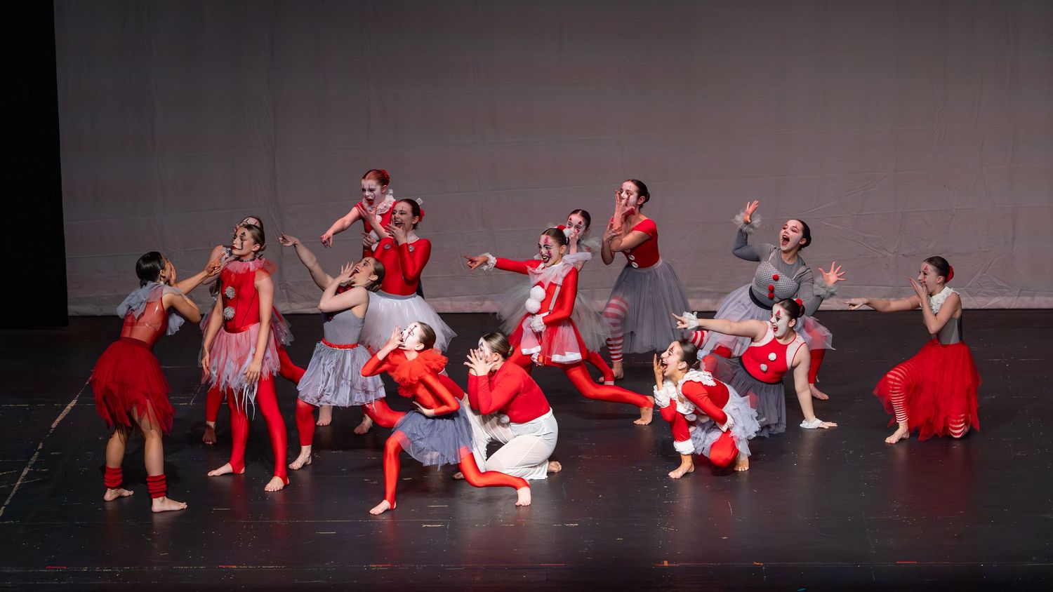Dancers in red and gray costumes perform a dramatic group ballet on a dark stage.