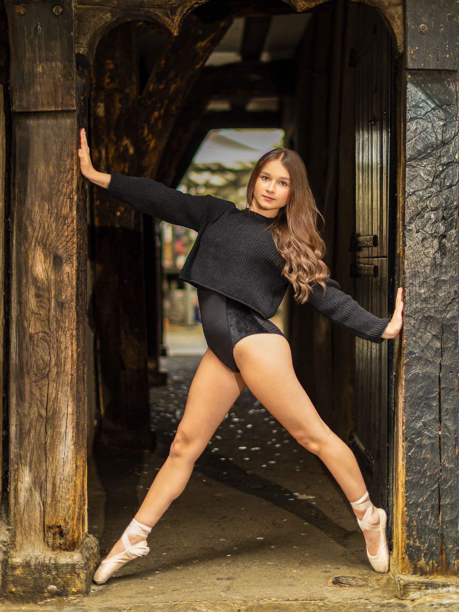 A ballet dancer in black leotard and pointe shoes strikes an elegant pose in a rustic wooden doorway.