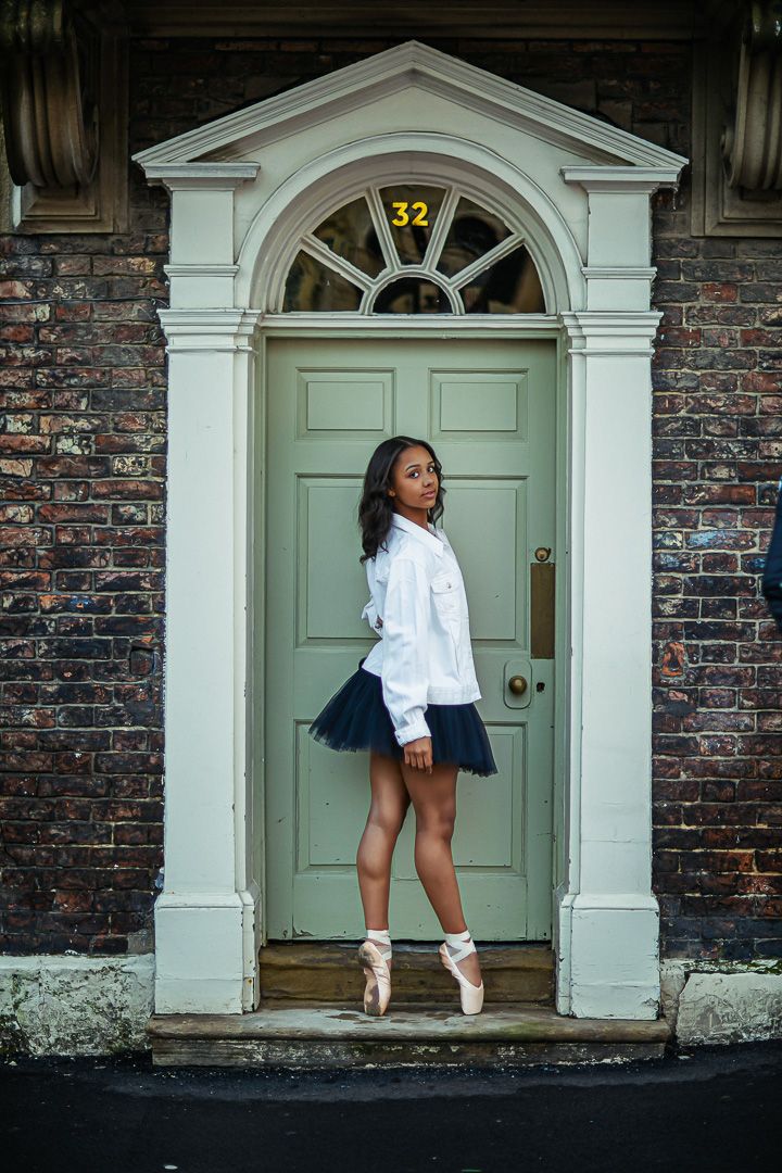 A young ballerina in white top and navy tutu poses en pointe before a sage green Georgian door numbered 32.