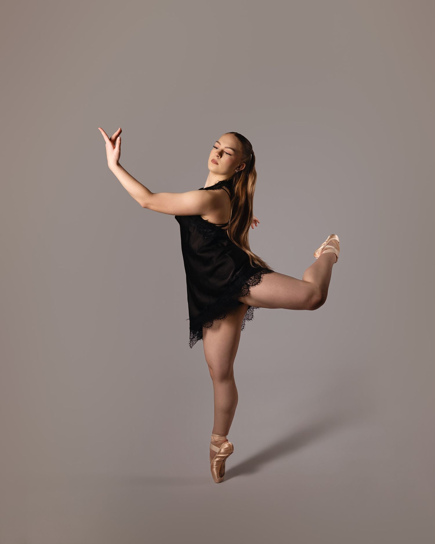 A ballet dancer in a black lace dress performs an arabesque pose on pointe shoes against a neutral gray background.