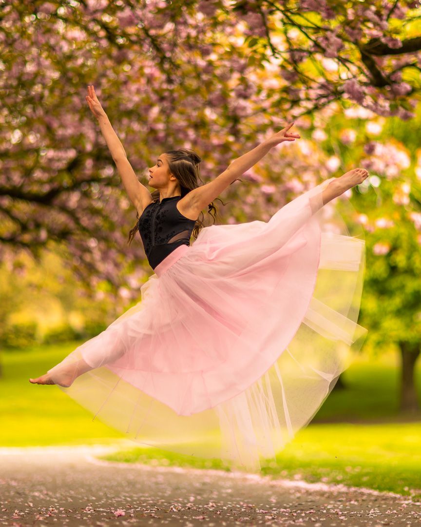 Flowing pink dress creates ethereal effect during dance leap under cherry blossoms.