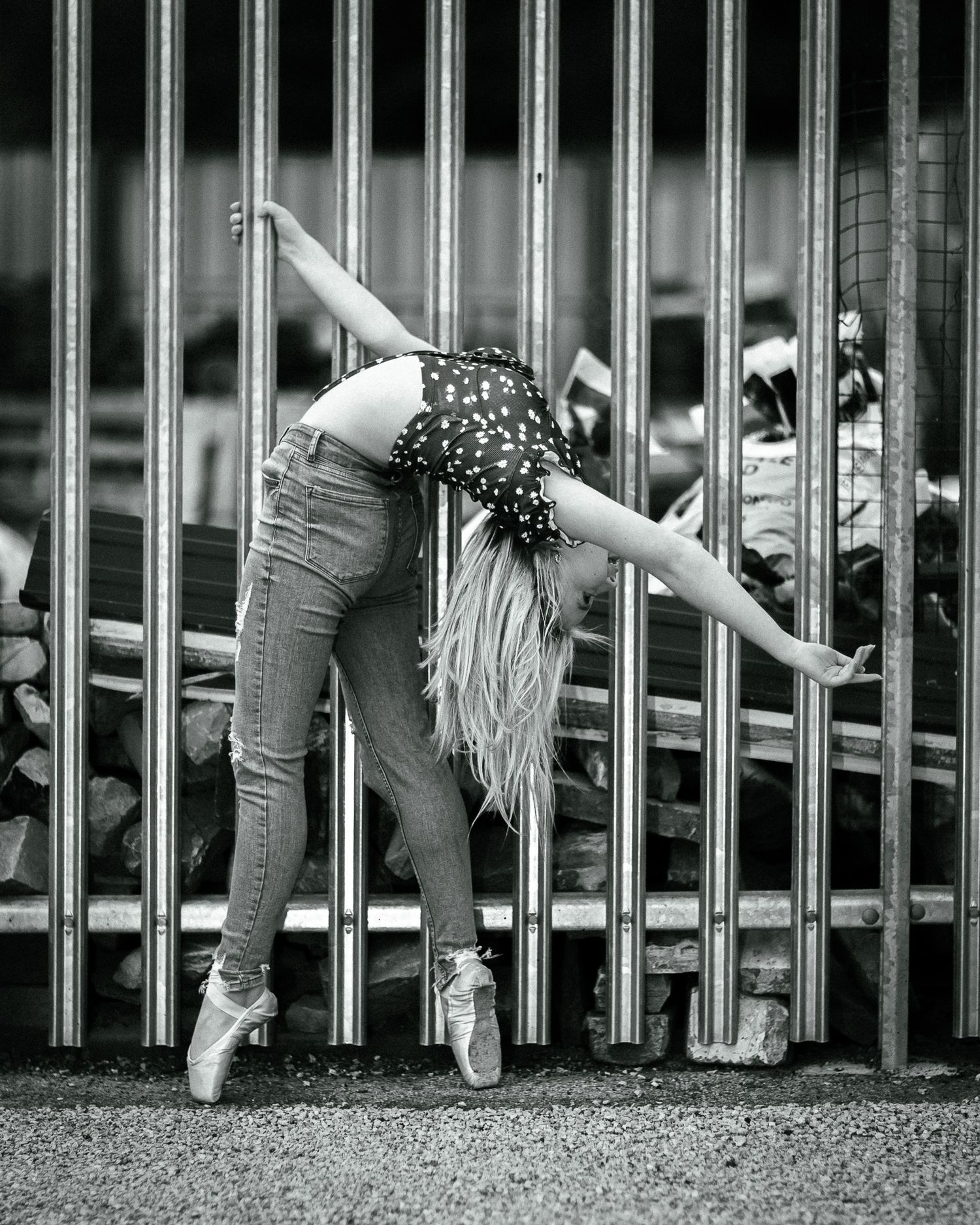 Black and white photo of a ballet dancer in jeans and pointe shoes bending gracefully against metal fence bars.