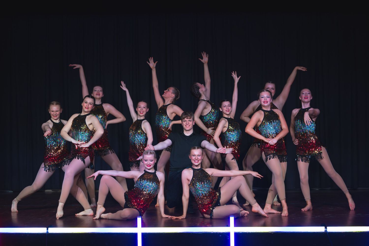 Group of dancers in sequined costumes performing on stage with dramatic lighting during a dance recital.