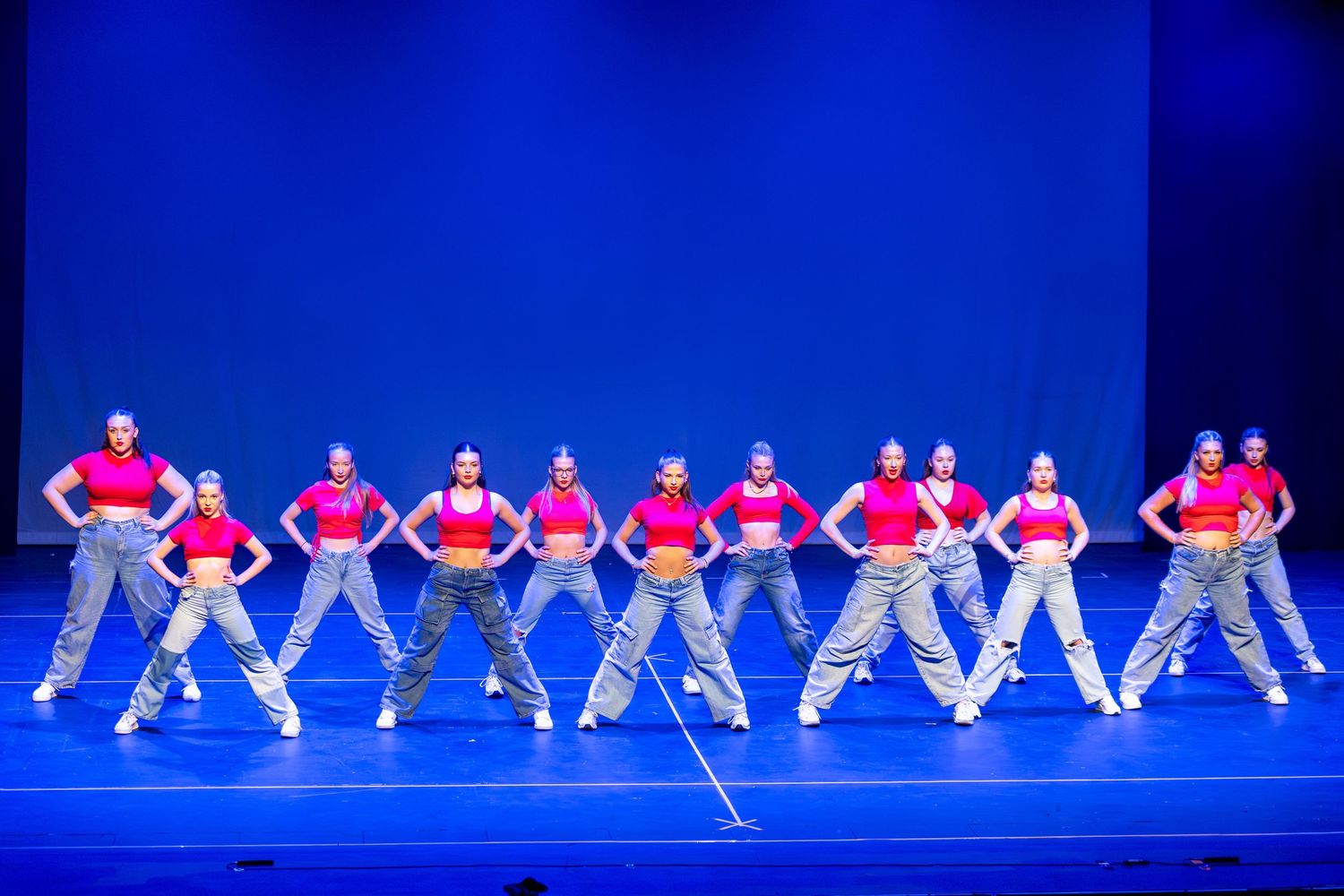 A dance troupe in matching pink tops and gray pants performs a synchronized routine on a blue-lit stage.