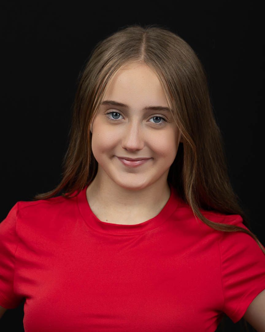 Young girl with long brown hair and blue eyes smiling, wearing a red t-shirt against a black background.