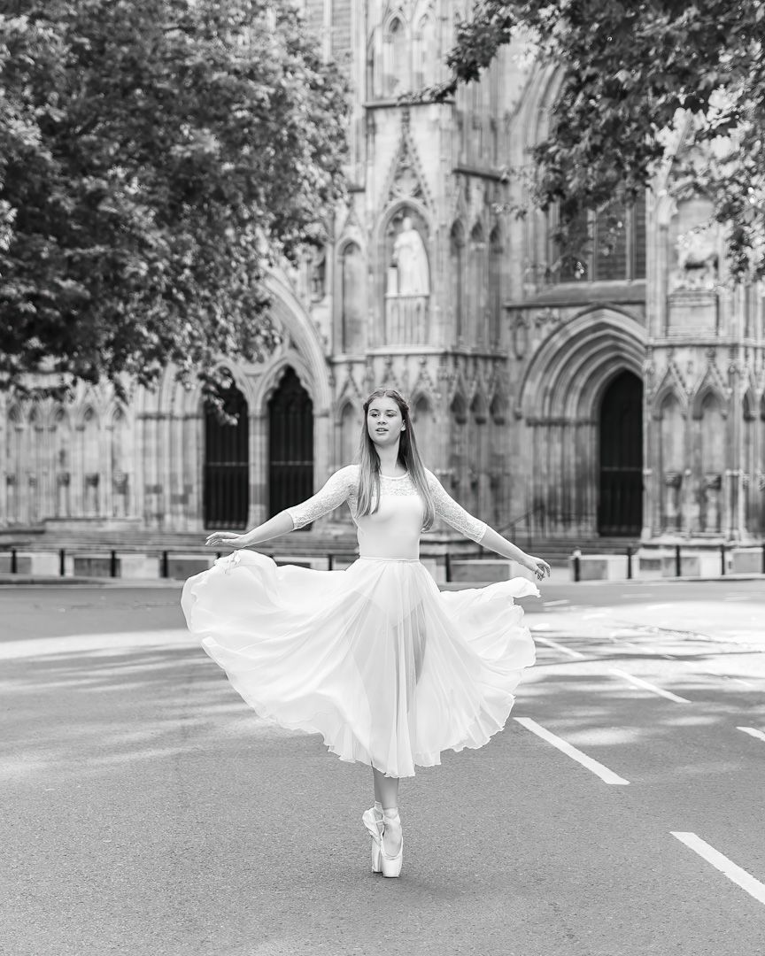 Black and white photo of a ballerina in a white dress dancing en pointe outside a Gothic cathedral.