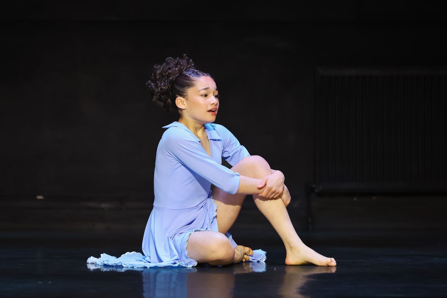 A female dancer sits gracefully on stage in a light blue costume, knees drawn up, performing a contemporary dance piece.