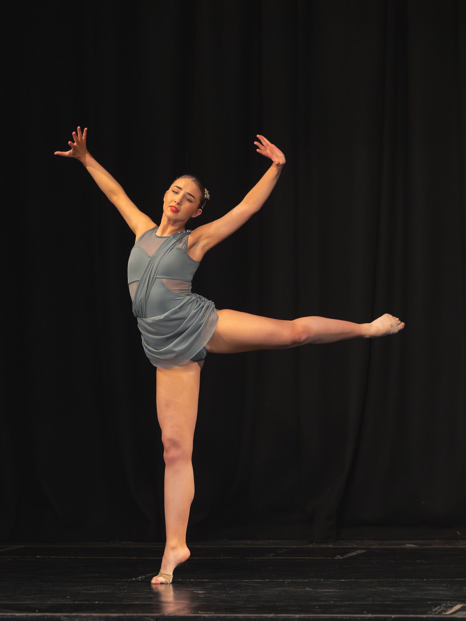A dancer performs an arabesque on stage in a gray costume, arms raised gracefully under black backdrop lighting.