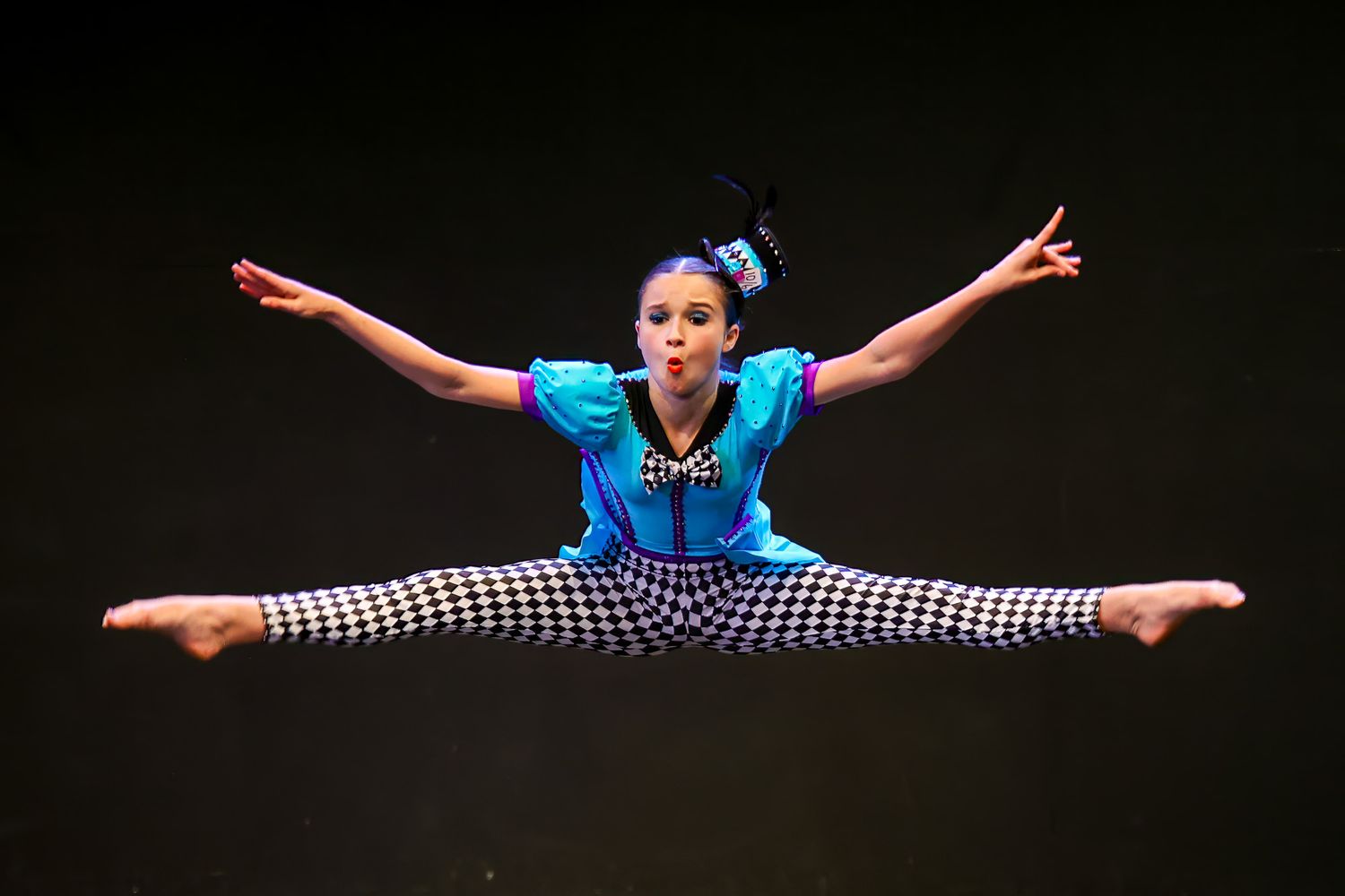 Dancer in mid-air split leap wearing blue top and hcheckered pants performing on stage.