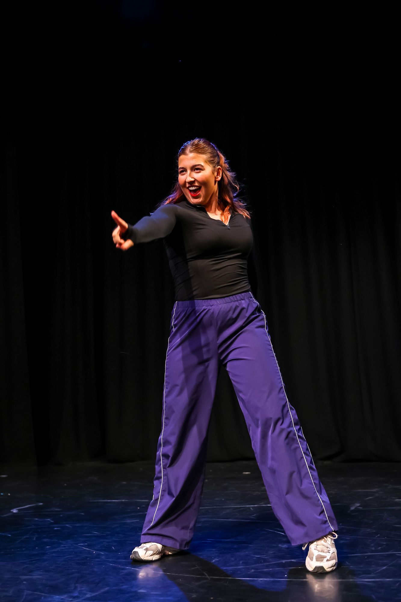 Young woman in black top and purple wide-leg pants dancing energetically on a dark stage.