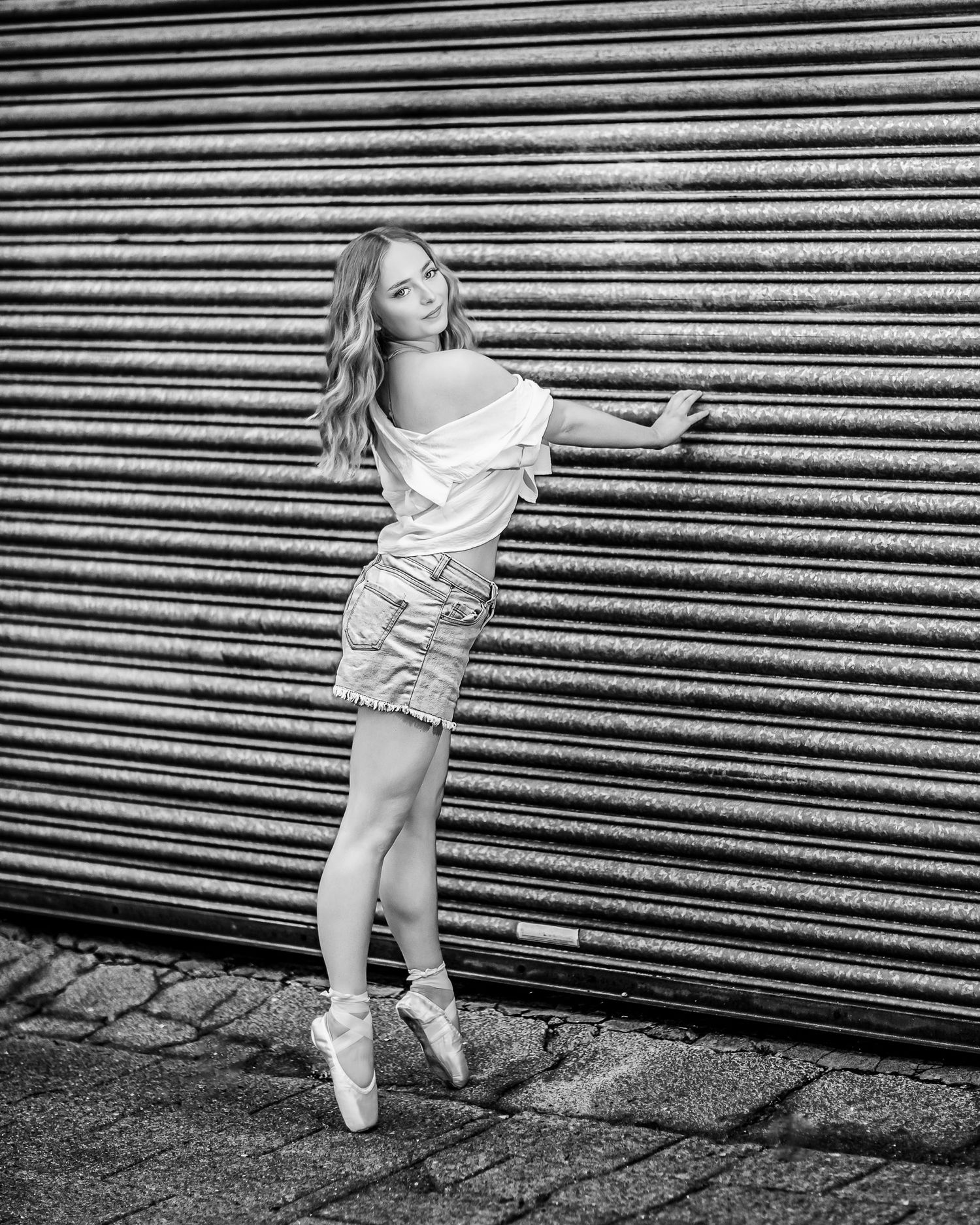 A young child in a white dress twirls playfully against a backdrop of horizontal metal shutters in black and white.