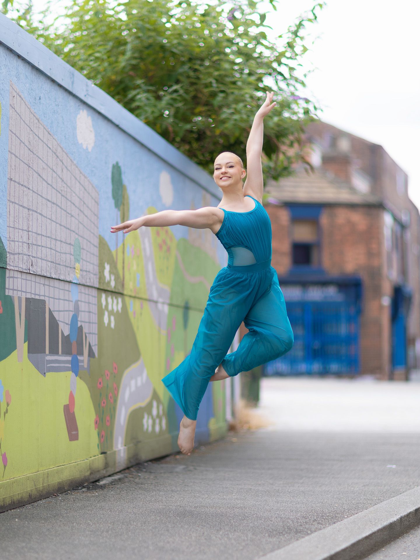 A dancer in a teal jumpsuit leaps gracefully in front of a colorful mural on an urban street.