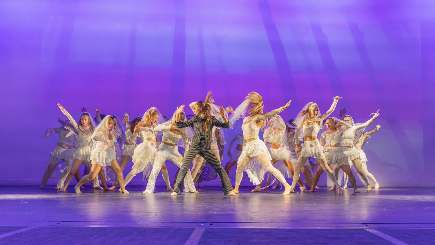Broadway dancers in white costumes perform on a purple-lit stage in a dramatic ensemble number.