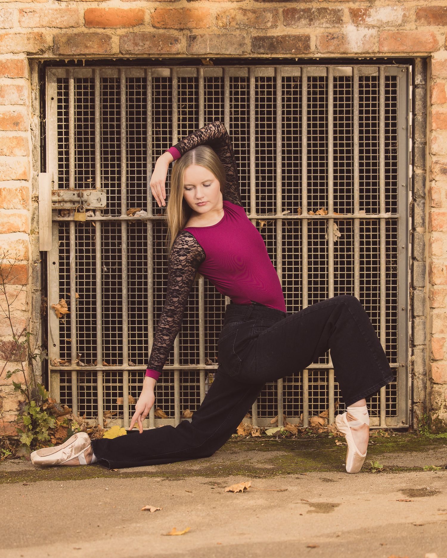 A ballet dancer in a magenta top and black pants poses gracefully in pointe shoes against a rustic brick wall.