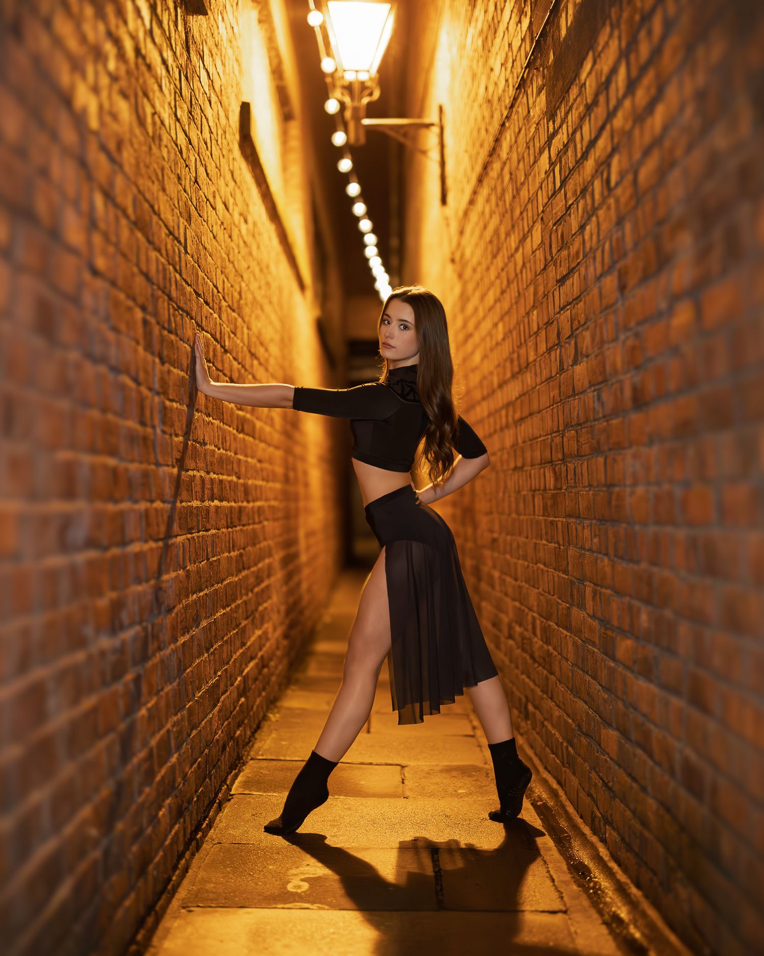 A woman in black crop top and pleated skirt poses dramatically in a narrow brick alley lit by warm golden lights.