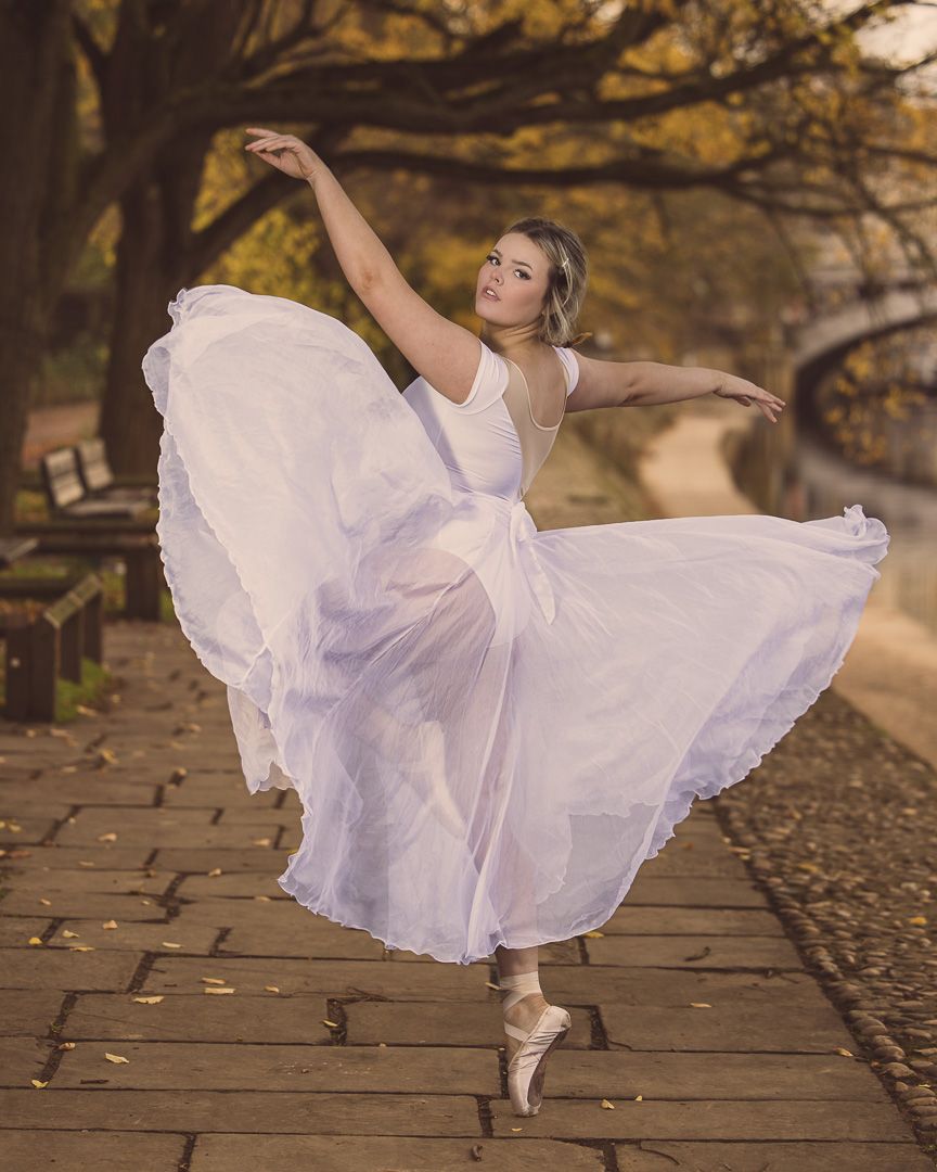 Ballerina twirls in flowing white tutu dress on stone pathway during fall season.