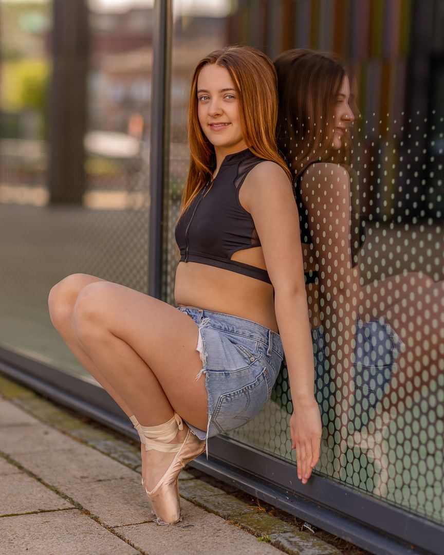 Dancer in denim shorts practices ballet poses along modern building wall.