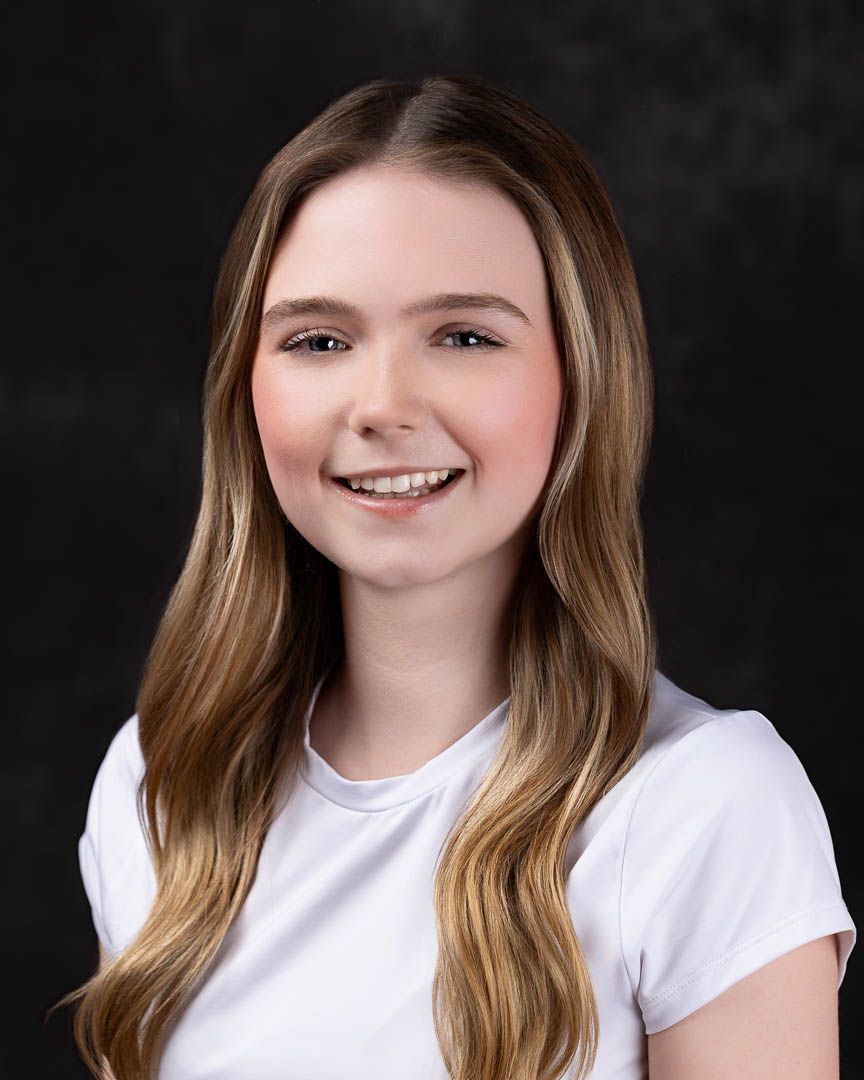 Young woman with long blonde hair smiling confidently in a white t-shirt against a dark background.