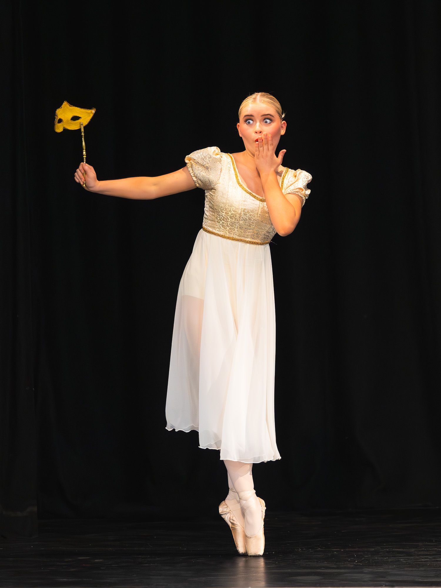Ballerina in white dress on pointe shoes holds gold masquerade mask, performing expressive dance on dark stage.