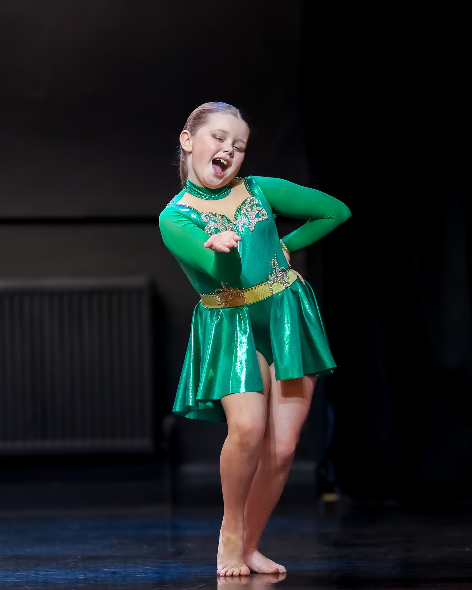 A young girl performs on stage in a shiny green dance costume with a gold belt, smiling expressively.
