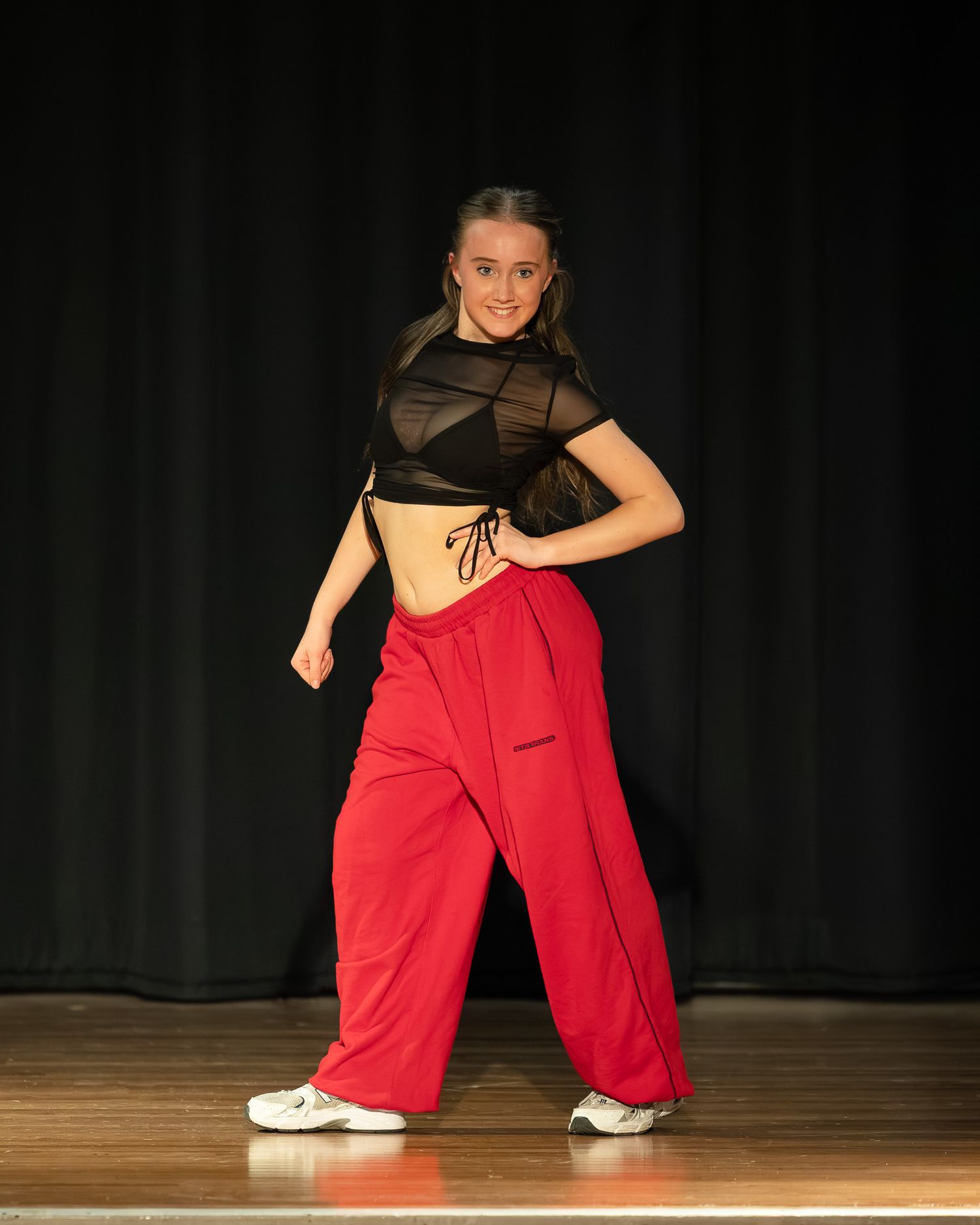 Young dancer performing on stage in red cargo pants and black mesh crop top under spotlight.