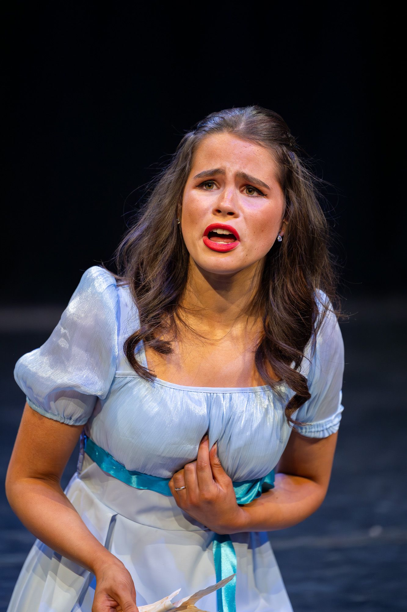 Young actress in blue dress performs emotional scene on stage, expressing distress with hand on chest.