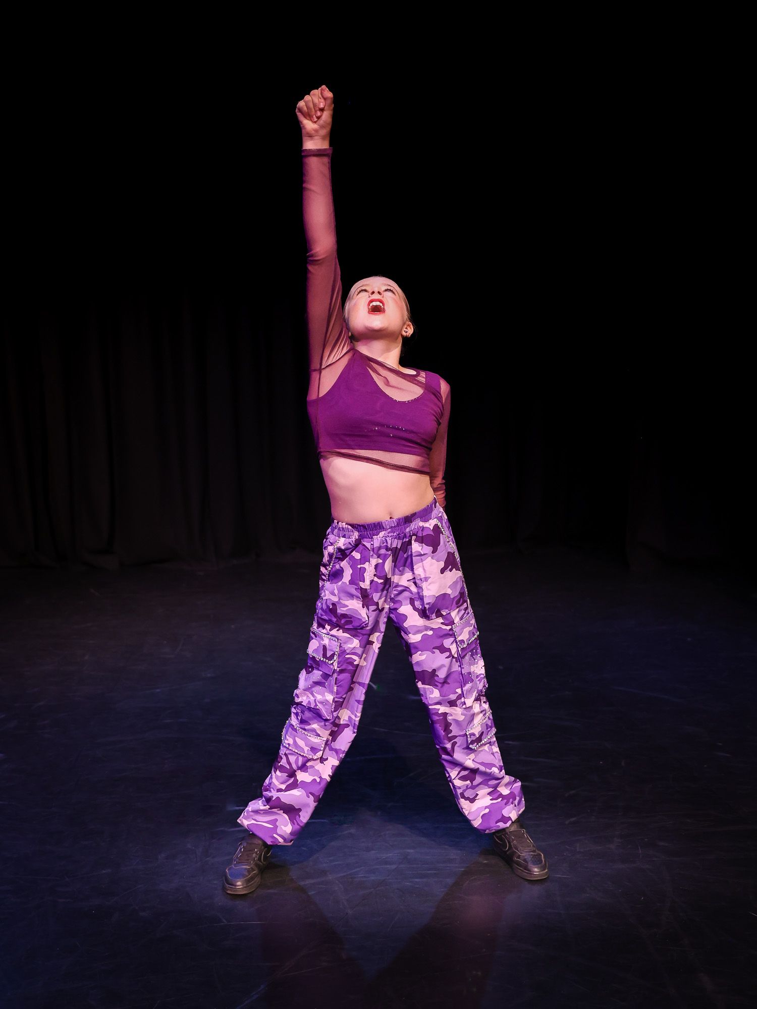 A dancer in purple camo pants and crop top performs on stage with one arm raised dramatically under spotlight.