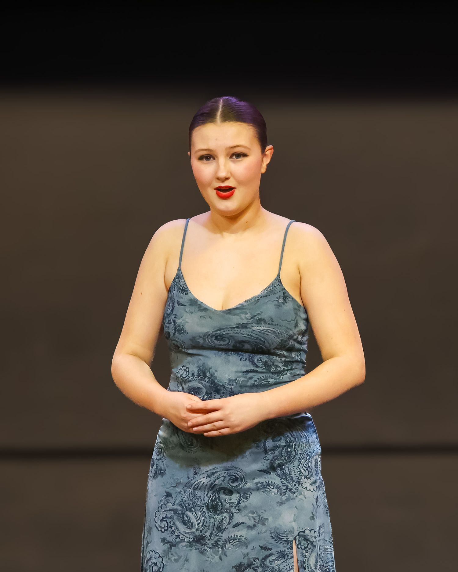 Young woman in blue floral satin slip dress performing on stage with red lipstick.