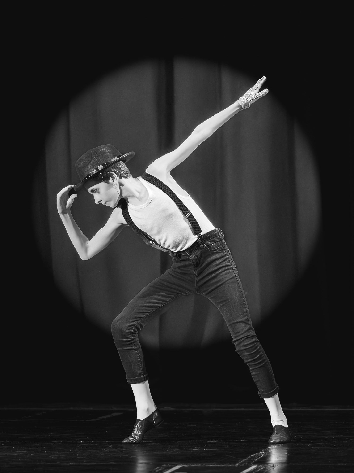 Black and white photo of a dancer in hat and suspenders striking a dramatic pose under a spotlight on stage.