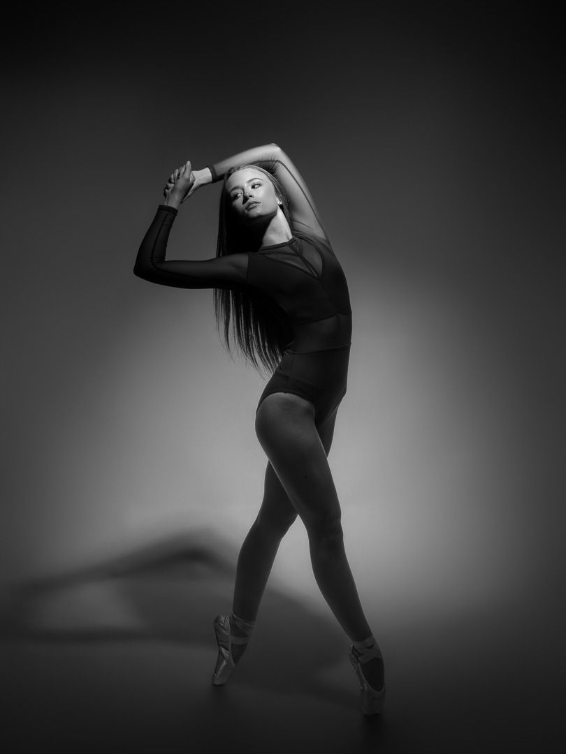 Black and white photo of a ballet dancer in a black leotard posing gracefully on pointe shoes in a studio.