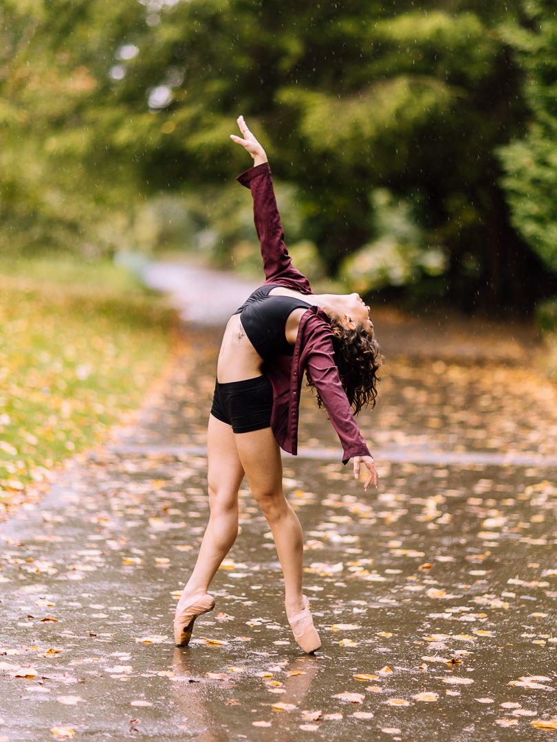 A ballet dancer in pointe shoes performs a graceful backbend in the rain on a wet path surrounded by autumn foliage.
