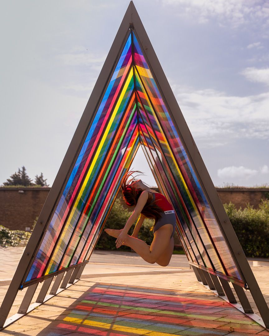 Dancer leaps through colorful rainbow-striped triangular art installation.