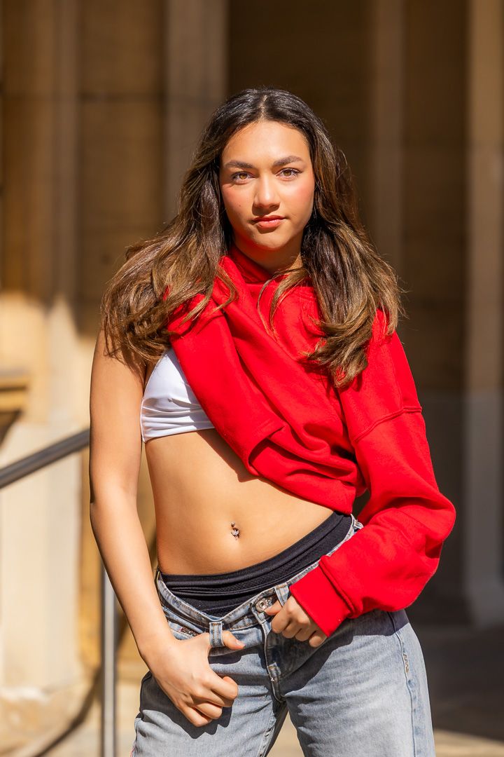 Urban dance photoshoot featuring red cropped hoodie and denim style against architectural backdrop.