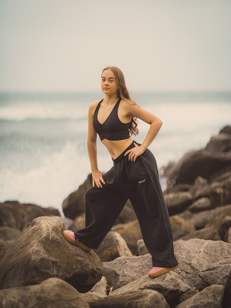 Athletic dancer poses on rocky coastline wearing black crop top and loose pants.
