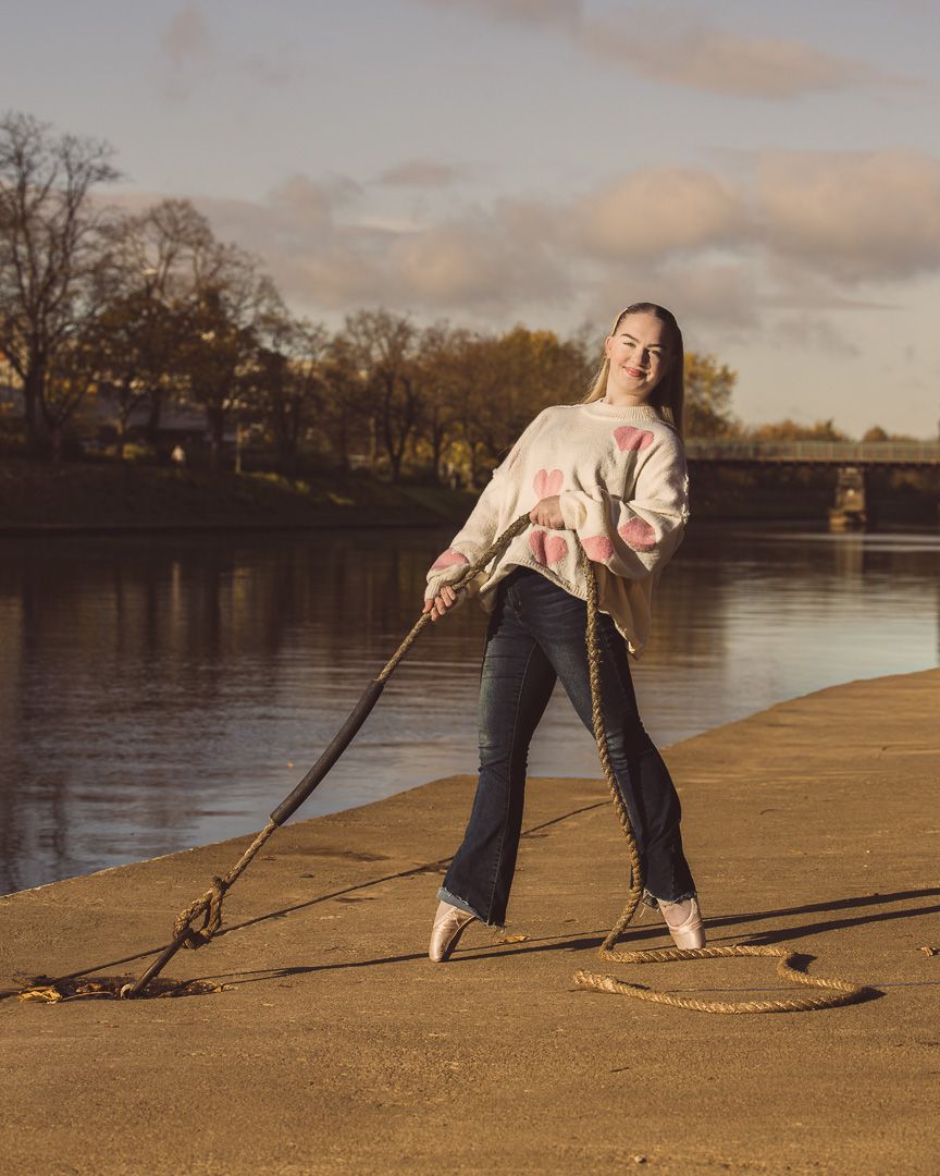 Dancer in pink sweater and jeans poses playfully with a dog leash by a river at sunset.