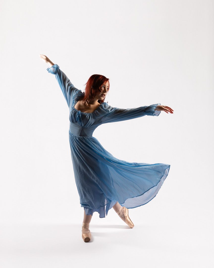Dancer in flowing blue dress performs graceful arabesque pose against white background.
