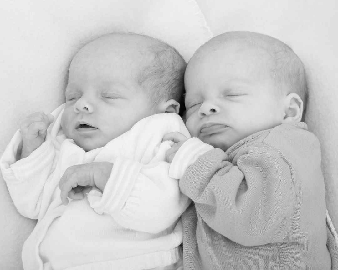 Black and white photo of two sleeping newborn babies lying side by side in matching outfits.