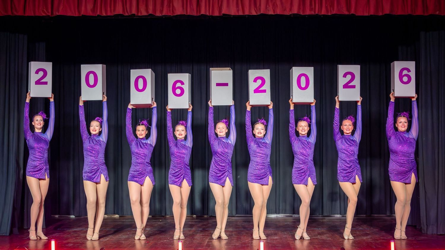 Eight dancers in purple costumes hold signs spelling 2006-2026 on stage.
