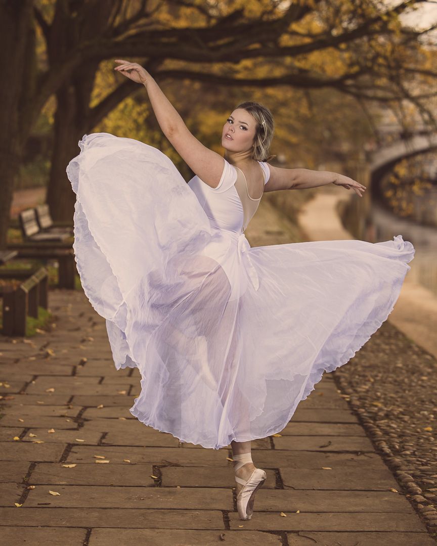 Ballerina in flowing white dress dancing en pointe on autumn park path with warm golden foliage backdrop.