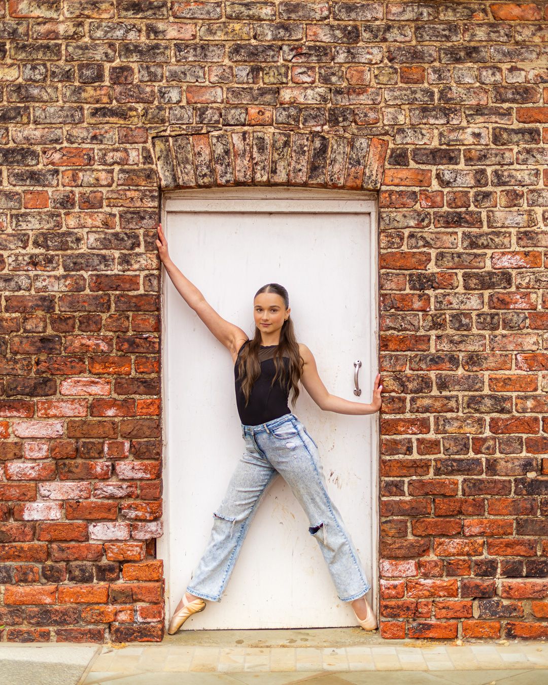 Young woman in black top and jeans posing in a white doorway framed by a rustic red brick wall.